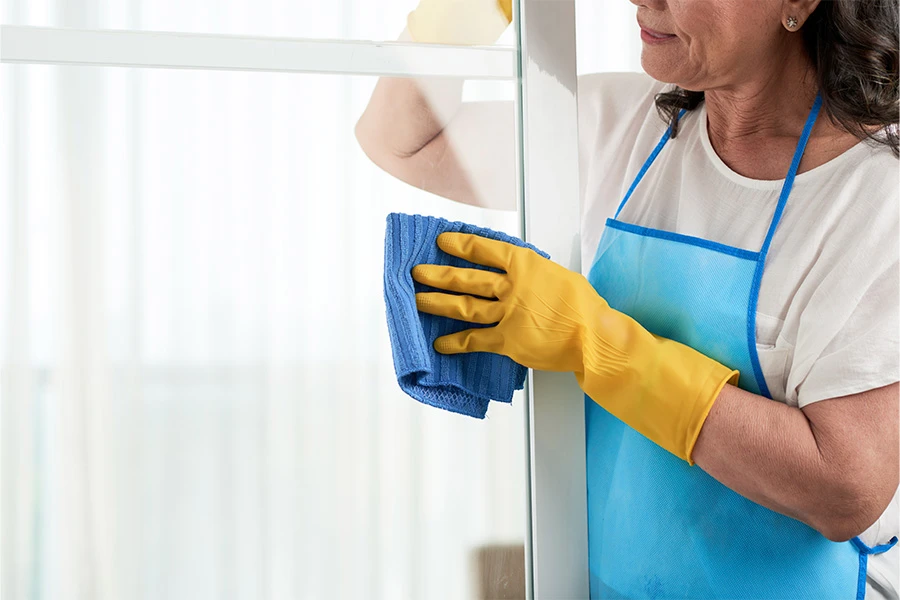 Woman in yellow gloves wiping a glass panel with a blue microfiber cloth.