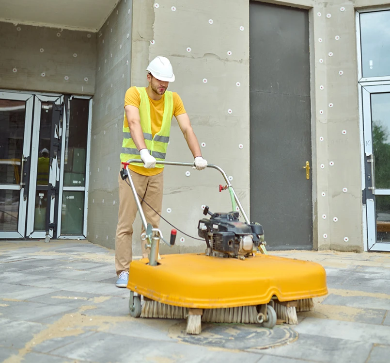 Worker in vest and hard hat using an outdoor floor sweeper on paving at a building site.