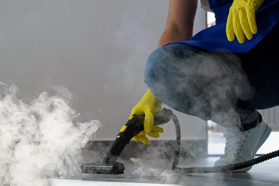 Person in yellow gloves using a handheld steam cleaner on a tile floor.