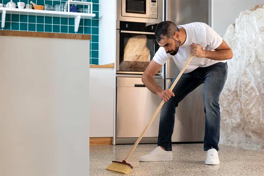 Man in a white shirt sweeping a kitchen floor with a broom.